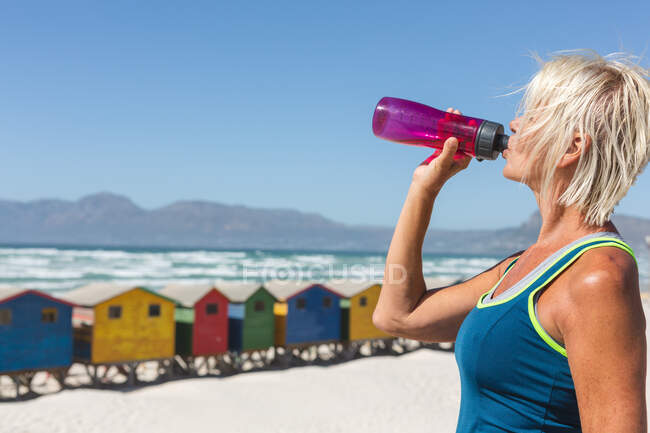 Senior Caucasian woman enjoying exercising on a beach on a sunny day, resting after running on the seashore and drinking water from a bottle with little colorful houses in the background. — Stock Photo