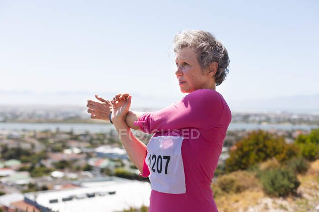 Mujer blanca mayor disfrutando del ejercicio en un día soleado, estirándose antes de correr, usando números y ropa deportiva rosa, con el cielo azul en el fondo. - foto de stock