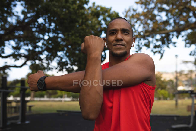 Hombre de raza mixta confiado usando ropa deportiva, ejercitándose en un parque, estirando sus brazos usando reloj inteligente y auriculares inalámbricos. Fitness fuerza estilo de vida saludable. - foto de stock