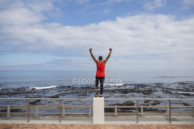 Rear view of disabled mixed race man with a prosthetic leg and running blade working out by the coast, standing on a fence looking out to sea with arms raised. Fitness disability healthy lifestyle. — Stock Photo