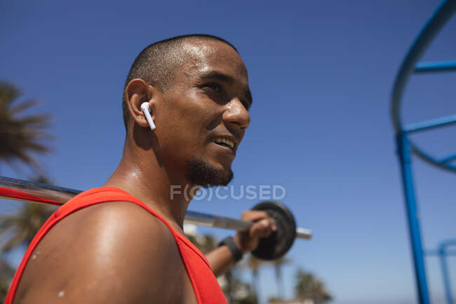 Hombre de raza mixta seguro con un entrenamiento en un gimnasio al aire libre bajo el sol, usando auriculares inalámbricos, sosteniendo pesas en una barra. Fitness discapacidad estilo de vida saludable. - foto de stock