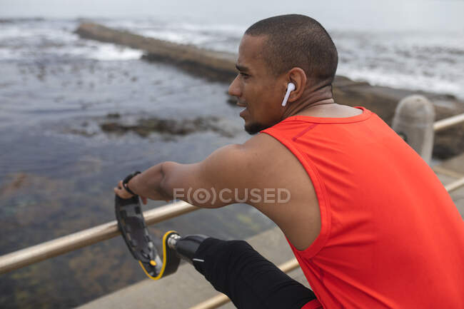 Disabled mixed race man with a prosthetic leg and running blade working out by the coast wearing wireless earphones, stretching with running blade up on a fence. Fitness disability healthy lifestyle. — Stock Photo