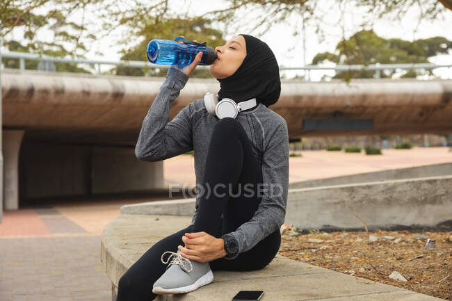 Fit gemischte Rennläuferin mit Hijab und Sportkleidung, die draußen in der Stadt trainiert, trinkt aus einer Wasserflasche, während sie mit Kopfhörern im Stadtpark Pause macht. Bewegung im städtischen Lebensstil. — Stockfoto