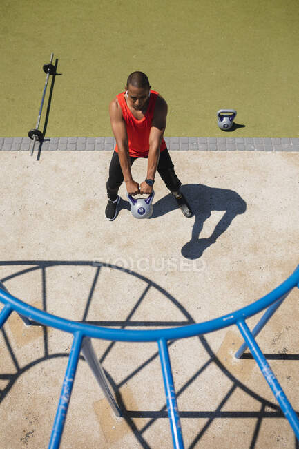 High angle view of disabled mixed race man with a prosthetic running blade working out at an outdoor gym wearing wireless earphones, swinging a kettlebell weight. Fitness disability healthy lifestyle. — Stock Photo