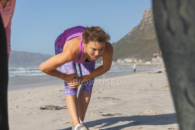 Donna caucasica con i capelli castani che si esercita su una spiaggia in una giornata di sole, pratica yoga e stretching con il mare sullo sfondo. — Foto stock