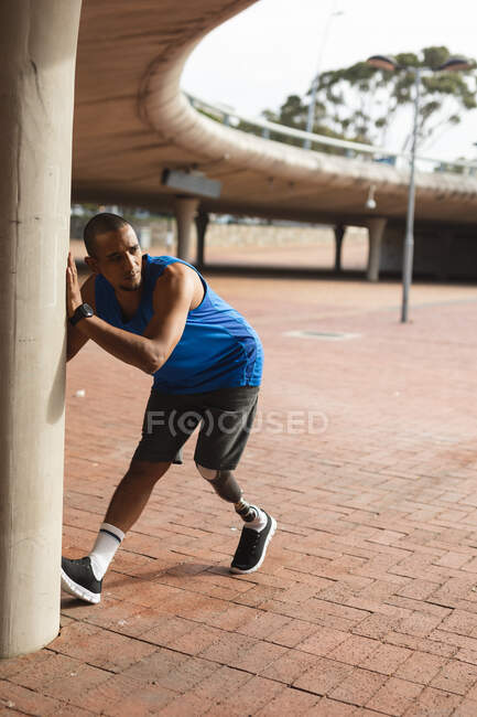 Hombre de raza mixta discapacitado con una pierna protésica, haciendo ejercicio en un parque urbano, apoyado contra la pared, estirando la pierna. Fitness discapacidad estilo de vida saludable. - foto de stock