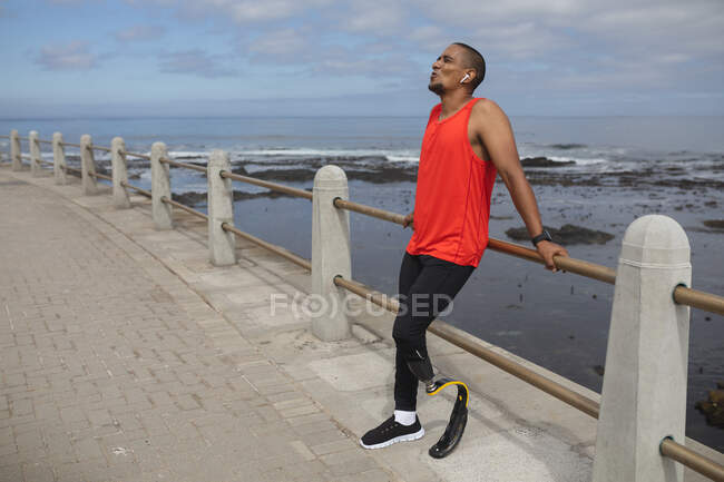Disabled mixed race man with a prosthetic leg and running blade working out by the coast wearing wireless earphones, taking a break leaning on a fence. Fitness disability healthy lifestyle. — Stock Photo