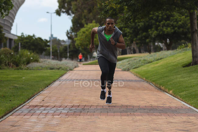 Uomo disabile di razza mista con una gamba protesica, che si allena in un parco urbano, indossando un top incappucciato che corre su un sentiero. Fitness disabilità stile di vita sano. — Foto stock