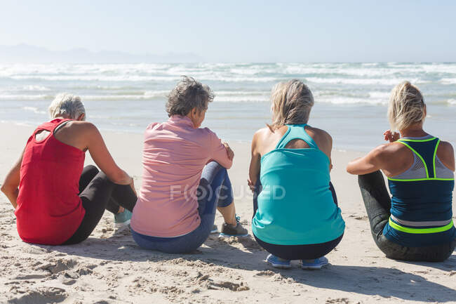 Eine Gruppe kaukasischer Freundinnen genießt an einem sonnigen Tag das Training am Strand, sitzt auf Sand und blickt auf das Meer. — Stockfoto
