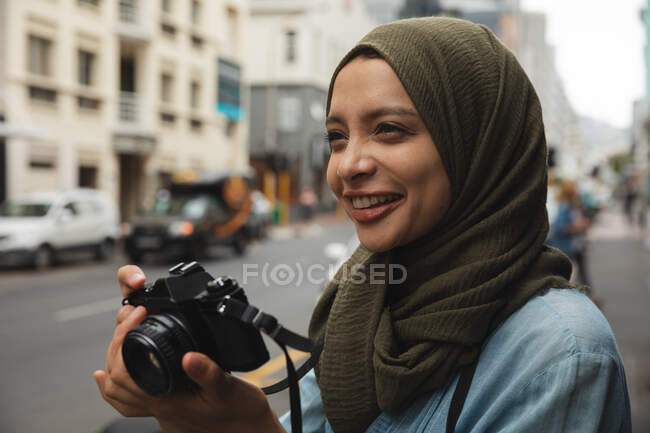 Mujer de raza mixta con hijab recorriendo la ciudad, sonriendo con la cámara digital tomando fotos. Turismo turismo moderno estilo de vida. - foto de stock