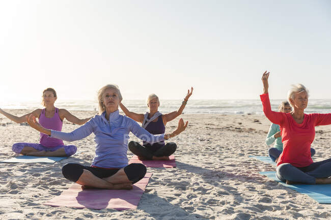 Группа кавказских подруг, занимающихся спортом на пляже в солнечный день, практикующих йогу, медитирующих в позе лотоса на заднем плане. — стоковое фото