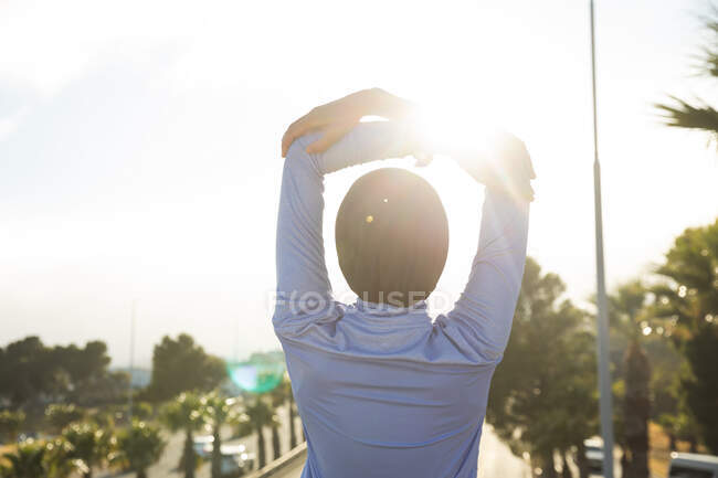 Rear view of fit mixed race woman wearing hijab and sportswear exercising outdoors in the city on a sunny day, stretching her arms on a footbridge. Urban lifestyle exercise. — Stock Photo
