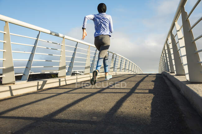 Rear view of fit mixed race woman wearing hijab and sportswear exercising outdoors in the city on a sunny day, running on a footbridge. Urban lifestyle exercise. — Stock Photo