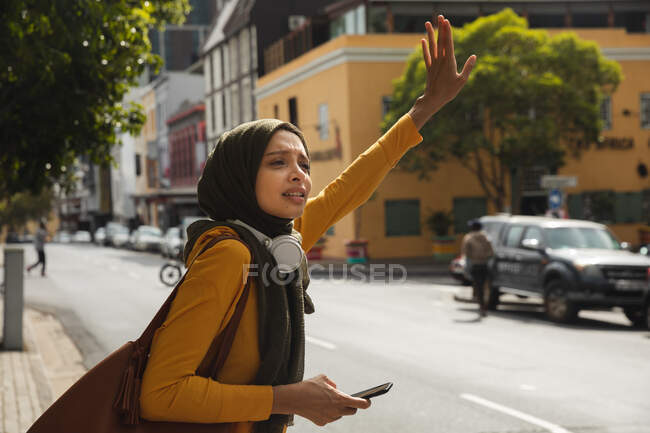 Donna di razza mista che indossa hijab e maglione giallo in giro per la città, fermando il taxi con il braccio in aria che tiene smartphone con cuffie wireless. Stile di vita moderno pendolare. — Foto stock