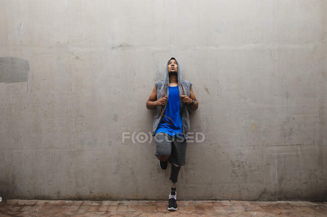 Homem de raça mista deficiente com uma perna protética, trabalhando em um parque urbano, vestindo top encapuzado encostado contra uma parede segurando pulando corda fazendo uma pausa. Incapacidade física estilo de vida saudável. — Fotografia de Stock
