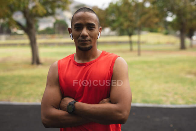 Portrait d'un homme de race mixte confiant portant des vêtements de sport, travaillant dans un parc, regardant la caméra porter des écouteurs sans fil et smartwatch. Fitness mode de vie sain. — Photo de stock