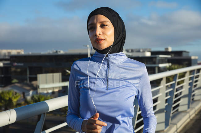 Fit mixed race woman wearing hijab and sportswear exercising outdoors in the city on a sunny day, running with earphones on a footbridge. Urban lifestyle exercise. — Stock Photo