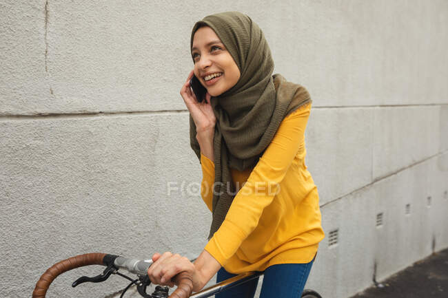 Жінка змішаної раси в хіджабі і жовтому джемпері і близько в дорозі в місто, розмовляючи на своєму смартфоні, посміхаючись сидячи на велосипеді. Комп'ютерний сучасний спосіб життя . — стокове фото