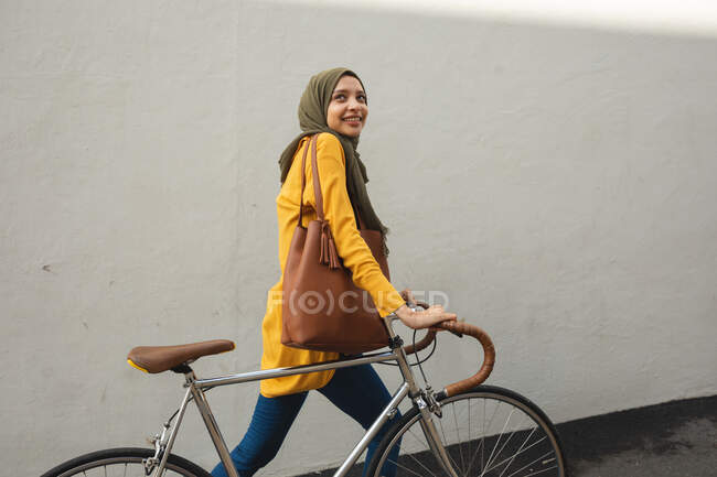 Жінка змішаної раси в хіджабі і жовтому джемпері і близько в дорозі в місто, посміхаючись ходити з велосипедом. Комп'ютерний сучасний спосіб життя . — стокове фото