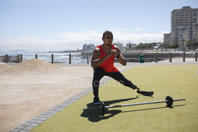 Behinderter Mischlingsrennfahrer mit Beinprothese und Laufklinge in einem Outdoor-Fitnessstudio an der Küste, mit drahtlosen Kopfhörern, streckenden Beinen und einer Langhantel neben ihm. Fitness Behinderung gesunder Lebensstil. — Stockfoto