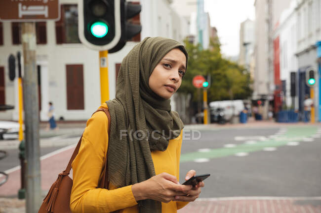 Mujer de raza mixta que usa hijab y jersey amarillo de ida y vuelta en la ciudad, sosteniendo su teléfono inteligente. Commuter estilo de vida moderno. - foto de stock