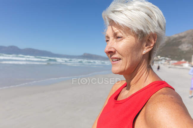 Старшая кавказская женщина наслаждается тренировками на пляже в солнечный день, стоя и любуясь видом на море на заднем плане. — стоковое фото