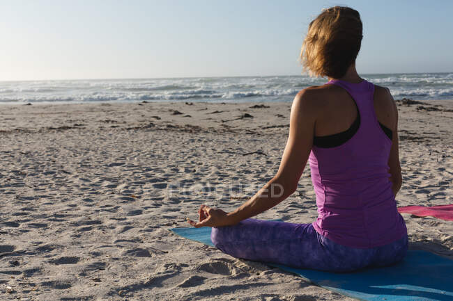 Caucasian woman with blond hair enjoying exercising on a beach on a sunny day, practicing yoga and meditating in lotus position, facing the sea. — Stock Photo