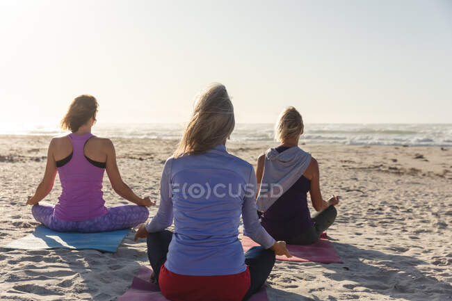 Группа кавказских подруг, занимающихся спортом на пляже в солнечный день, практикующих йогу, медитирующих в позе лотоса, стоящих лицом к морю. — стоковое фото