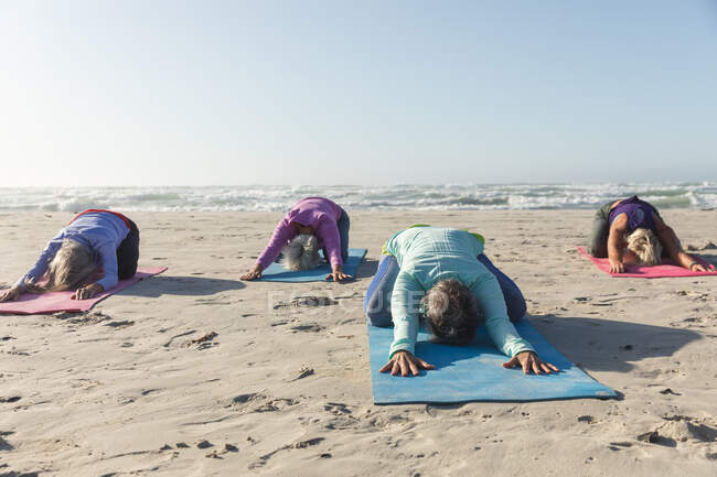 Группа кавказских подруг, занимающихся спортом на пляже в солнечный день, практикующих йогу и сидящих в позиции йоги. — стоковое фото