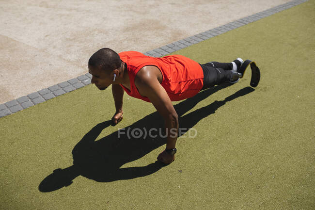 Disabled mixed race man with a prosthetic leg and running blade exercising at an outdoor gym by the coast wearing wireless earphones and doing press ups. Fitness disability healthy lifestyle. — Stock Photo