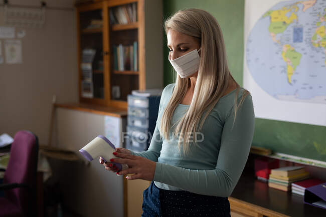 Professeure caucasienne portant un masque facial se préparant à mesurer la température dans une école primaire. Enseignement primaire distanciation sociale sécurité sanitaire pendant la pandémie de coronavirus Covid19. — Photo de stock