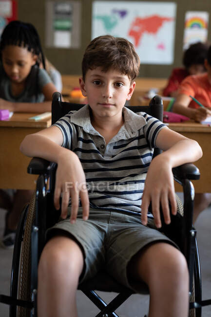 Retrato de un niño caucásico incapacitado sentado en su silla de ruedas en el aula durante la lección. Educación primaria distanciamiento social seguridad sanitaria durante la pandemia del Coronavirus Covid19. - foto de stock