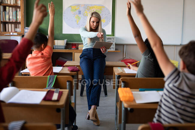 Group of multi ethnic kids sitting on their desk during the lesson with a Caucasian female teacher. Primary education social distancing health safety during Covid19 Coronavirus pandemic. — Stock Photo