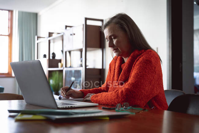 Femme caucasienne profitant du temps à la maison, de la distance sociale et de l'isolement personnel en quarantaine, assise à table, utilisant un ordinateur portable, prenant des notes. — Photo de stock