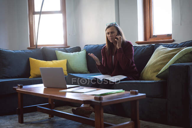 Кавказька жінка, яка проводить час удома, спілкується на відстані і самотності в карантині, сидить на дивані в вітальні, користується ноутбуком, розмовляє по телефону.. — стокове фото