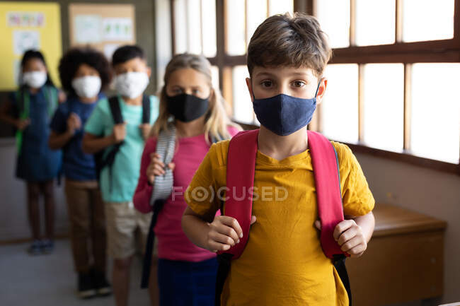 Multiethnische Gruppe von Grundschulkindern, die in die Kamera schauen und in der Schulaula Gesichtsmasken tragen. Grundschulbildung soziale Distanzierung der Gesundheitssicherheit während der Covid19 Coronavirus-Pandemie. — Stockfoto
