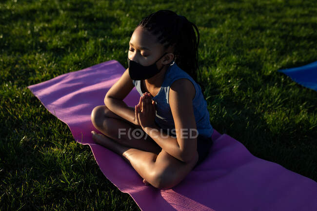 Mixed Race Girl mit Gesichtsmaske beim Yoga im Schulgarten. Grundschulbildung soziale Distanzierung der Gesundheitssicherheit während der Covid19 Coronavirus-Pandemie. — Stockfoto