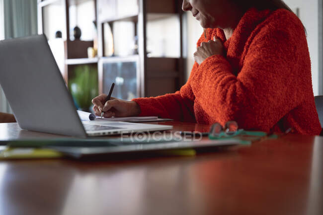 Donna caucasica godendo del tempo a casa, distanza sociale e auto isolamento in isolamento quarantena, seduto al tavolo, utilizzando un computer portatile e prendendo appunti. — Foto stock