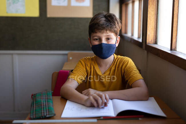 Retrato de un chico caucásico sentado en el escritorio con mascarilla en el aula. Educación primaria distanciamiento social seguridad sanitaria durante la pandemia del Coronavirus Covid19. - foto de stock