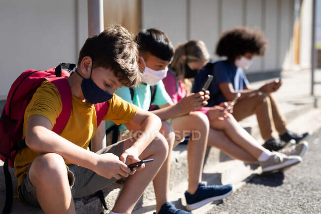 Grupo multiétnico de niños de la escuela primaria que usan máscaras faciales usando teléfonos inteligentes mientras están sentados juntos. Educación primaria distanciamiento social seguridad sanitaria durante la pandemia del Coronavirus Covid19 - foto de stock