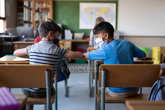 Two multi ethnic boys wearing face masks greeting each other by touching elbows at school. Primary education social distancing health safety during Covid19 Coronavirus pandemic. — Stock Photo