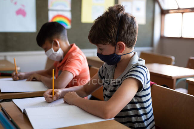 Zwei multiethnische Jungen sitzen im Klassenzimmer an Schreibtischen und tragen Gesichtsmasken. Grundschulbildung soziale Distanzierung der Gesundheitssicherheit während der Covid19 Coronavirus-Pandemie. — Stockfoto