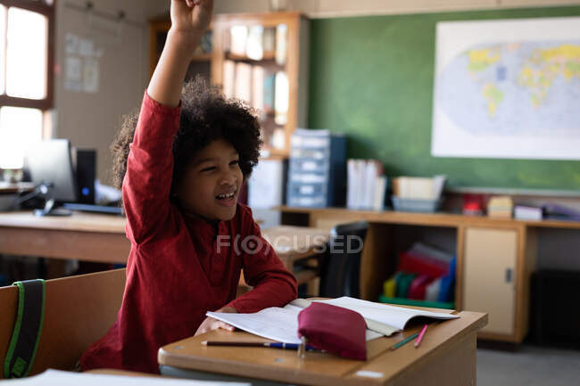 Niño de raza mixta levantando la mano mientras está sentado en su escritorio en la escuela. Educación primaria distanciamiento social seguridad sanitaria durante la pandemia del Coronavirus Covid19. - foto de stock