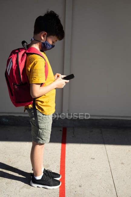 Caucasian boy wearing face mask, using his smartphone, maintaining social distancing at school. Primary education social distancing health safety during Covid19 Coronavirus pandemic. — Stock Photo