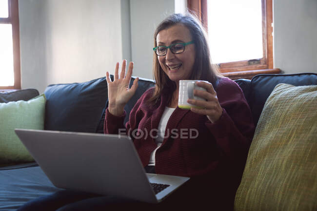 Femme caucasienne appréciant le temps à la maison, la distance sociale et l'isolement personnel en quarantaine verrouillage, assis sur le canapé dans le salon, à l'aide d'un ordinateur portable, agitant pendant l'appel vidéo. — Photo de stock