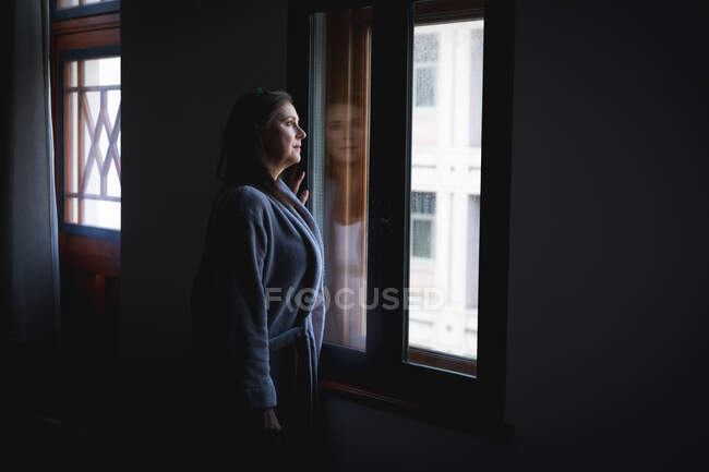 Mujer caucásica con cabello largo y oscuro disfrutando del tiempo en casa, distanciamiento social y auto aislamiento en cuarentena, de pie y mirando por la ventana. - foto de stock