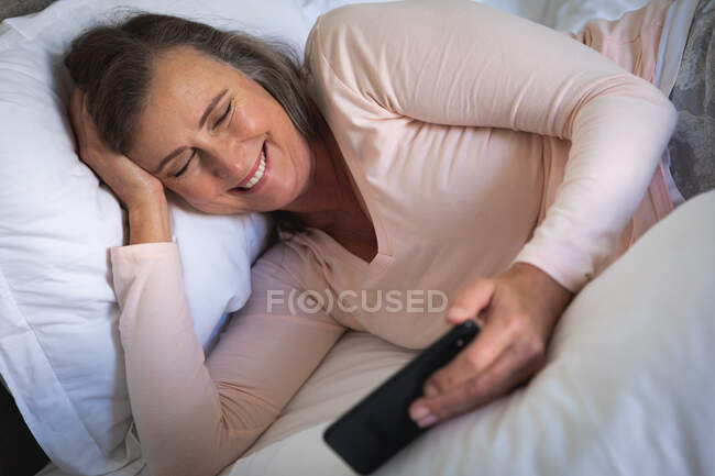 Кавказька жінка, яка проводить час удома, спілкується на відстані і самоізолюється в карантині, лежить у ліжку в спальні, користується смартфоном, посміхається. — стокове фото