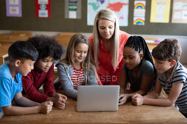 Insegnante caucasica femminile e gruppo multietnico di bambini che utilizzano il computer portatile durante la lezione. Istruzione primaria distanza sociale sicurezza sanitaria durante la pandemia di Covid19 Coronavirus. — Foto stock