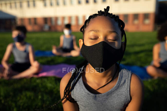 Mixed Race Girl mit Gesichtsmaske beim Yoga im Schulgarten. Grundschulbildung soziale Distanzierung der Gesundheitssicherheit während der Covid19 Coronavirus-Pandemie. — Stockfoto