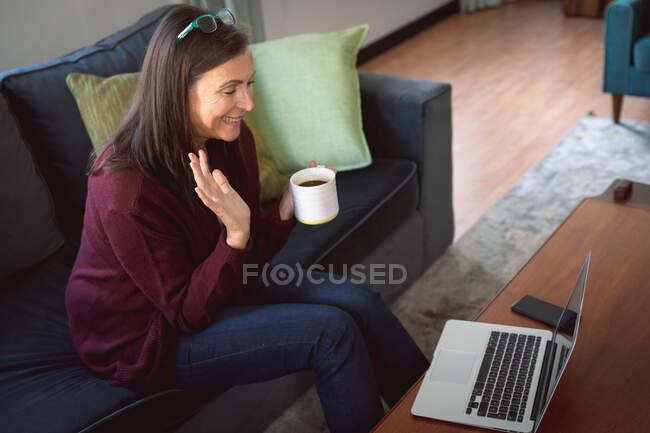 Caucasian woman enjoying time at home, social distancing and self isolation in quarantine lockdown, sitting on sofa in sitting room, using a laptop, waving during video call. — Stock Photo